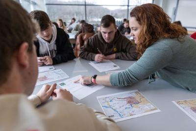 Four students collaborating across a table: tabletop is strewn with papers and students are leaning in to look and discuss them