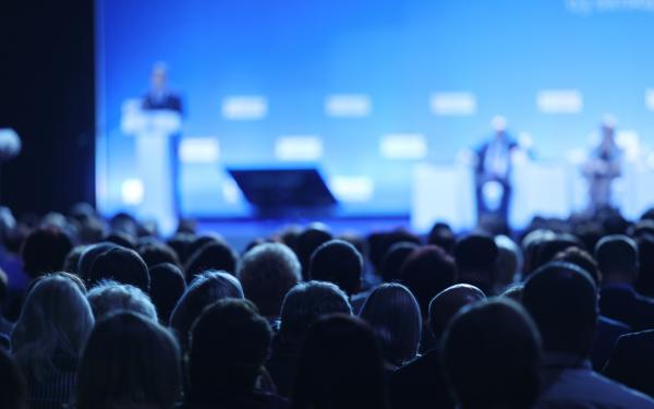 Lecture hall, with the backs of listerners' heads in the foreground and a out of focus stage in the background