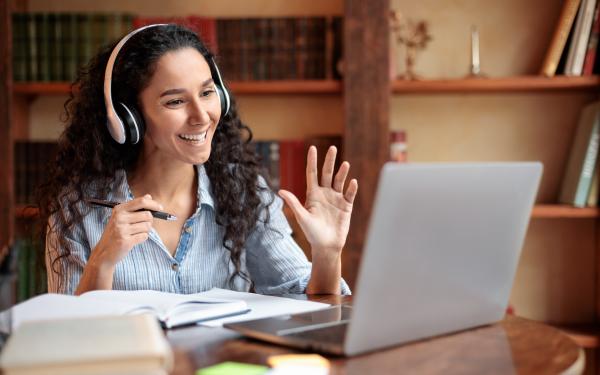 Student at desk with headphones and laptop, waiving to the computer screen.