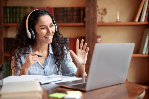 Student at desk with headphones and laptop, waiving to the computer screen.